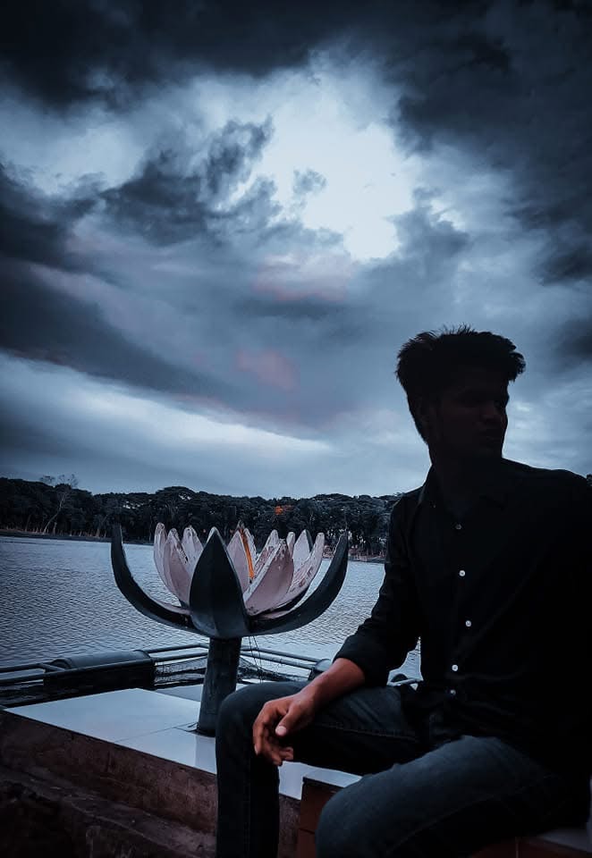 A seated figure silhouetted beside a river beneath dark clouds